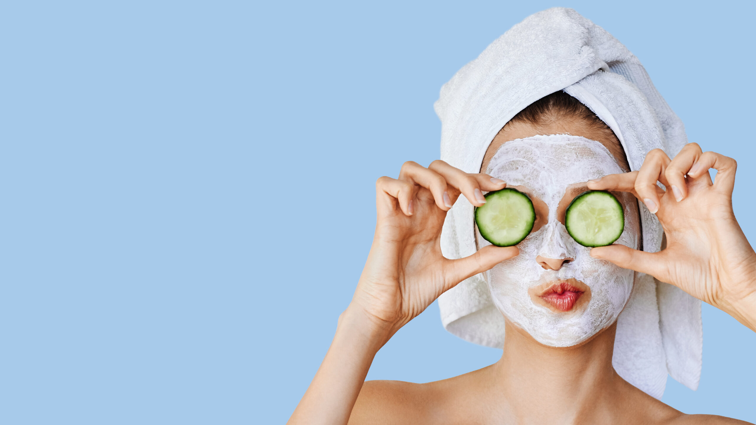 Beautiful young woman with facial mask on her face holding slices of fresh cucumber. Skin care and treatment, spa, natural beauty and cosmetology concept, over blue background with copy space